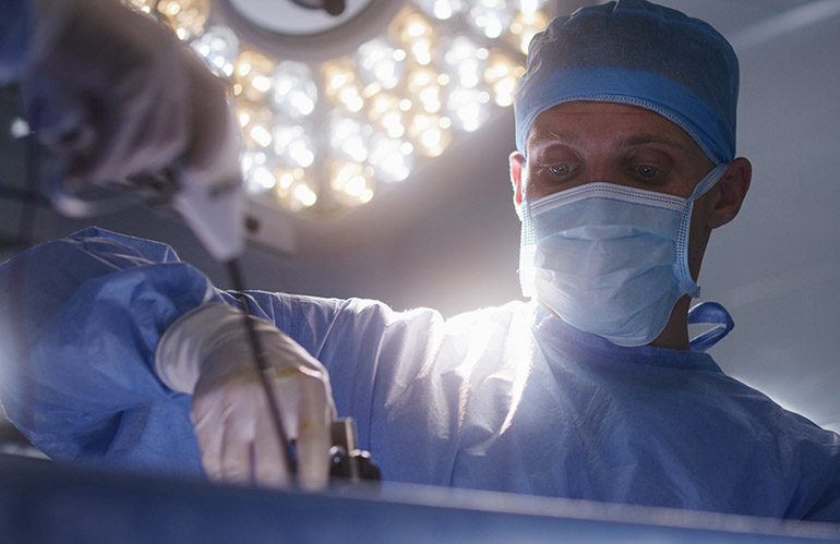 Close up of adult male medic assisting surgeon during surgery. Professional doctors operate patient using laparoscopy instruments in modern operating room. Medical staff at work in medical center.